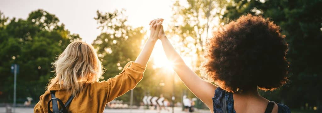 Couple of young women from the back, holding hands with arms raised and they walk in the street at sunset Two millennials are happy